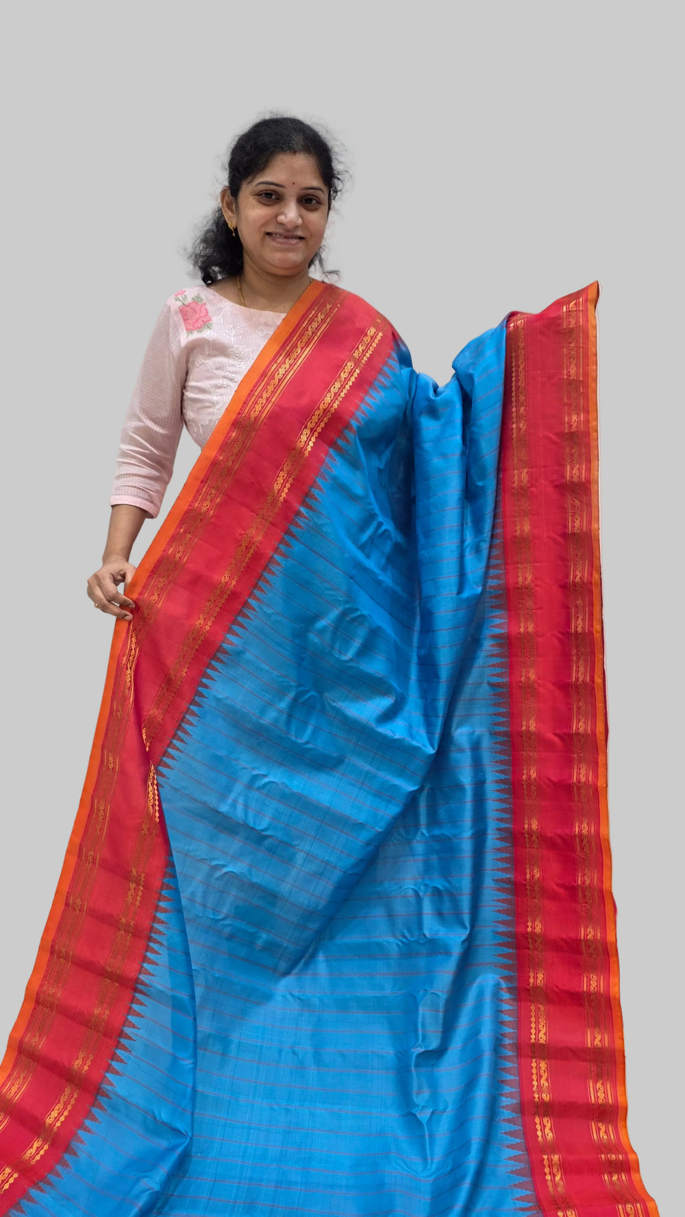 A woman modeling a vibrant blue and red silk saree with gold zari patterns, holding it up against a plain indoor wall.