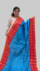 A woman modeling a vibrant blue and red silk saree with gold zari patterns, holding it up against a plain indoor wall.